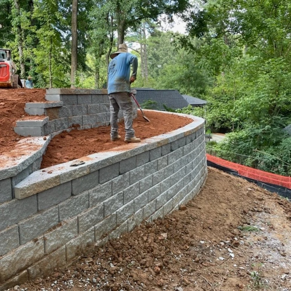 Contractor backfilling curved segmental block retaining wall on sloped yard in Metro Atlanta